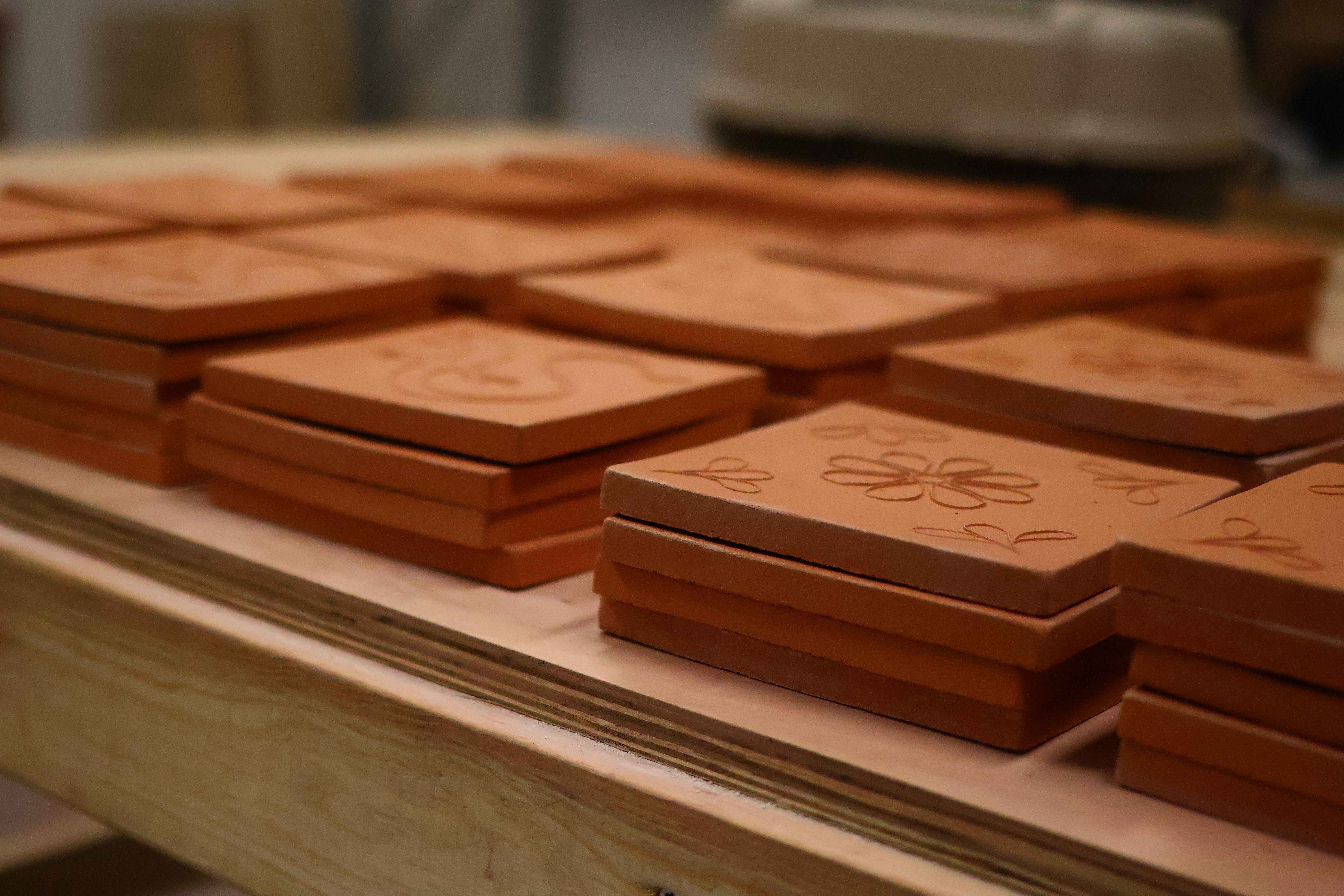 Stacks of raw terracotta tiles drying on a studio shelf, several carved with floral and scroll designs